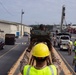 A military or industrial operation is underway in what appears to be a base or depot facility. A person wearing a yellow hard hat and high-visibility vest is directing a green armored military vehicle (likely a Stryker or similar wheeled armored personnel carrier) along a wooden ramp or track. Multiple workers in safety gear and a crane are visible in the background, suggesting a vehicle loading or positioning operation.