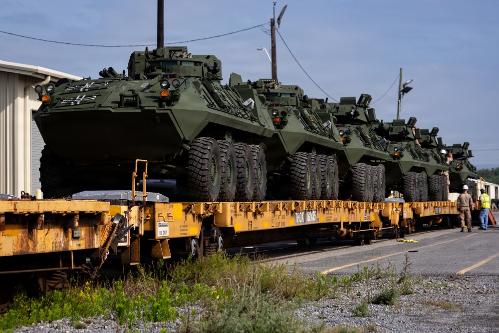 Multiple olive-drab military armored wheeled vehicles, consistent with Stryker-type infantry carrier vehicles, are loaded on yellow flatcar rail cars for transport. The vehicles are secured on what appear to be TPDX-marked military flatcars at a rail yard or military installation. Personnel in high-visibility vests and military uniforms are visible in the background, overseeing the loading or inspection process.