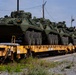 Multiple olive-drab military armored wheeled vehicles, consistent with Stryker-type infantry carrier vehicles, are loaded on yellow flatcar rail cars for transport. The vehicles are secured on what appear to be TPDX-marked military flatcars at a rail yard or military installation. Personnel in high-visibility vests and military uniforms are visible in the background, overseeing the loading or inspection process.