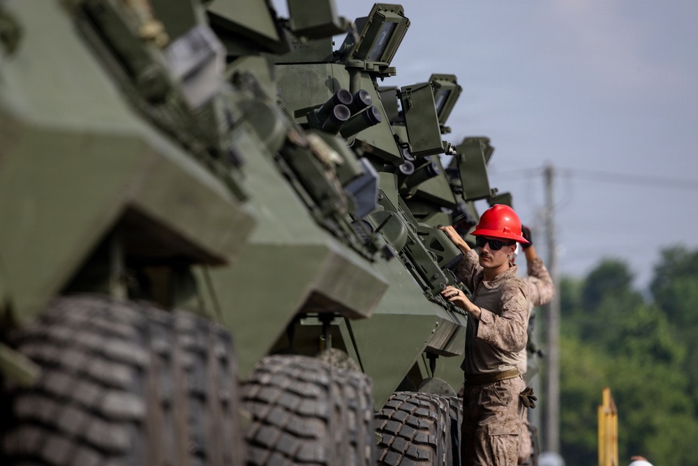 A military service member wearing a red hard hat and camouflage uniform is inspecting or performing maintenance on a row of olive-drab armored wheeled vehicles. The vehicles are lined up in formation, likely at a military installation or staging area. The scene suggests a pre-deployment inspection, equipment check, or acceptance procedure.