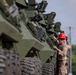 A military service member wearing a red hard hat and camouflage uniform is inspecting or performing maintenance on a row of olive-drab armored wheeled vehicles. The vehicles are lined up in formation, likely at a military installation or staging area. The scene suggests a pre-deployment inspection, equipment check, or acceptance procedure.
