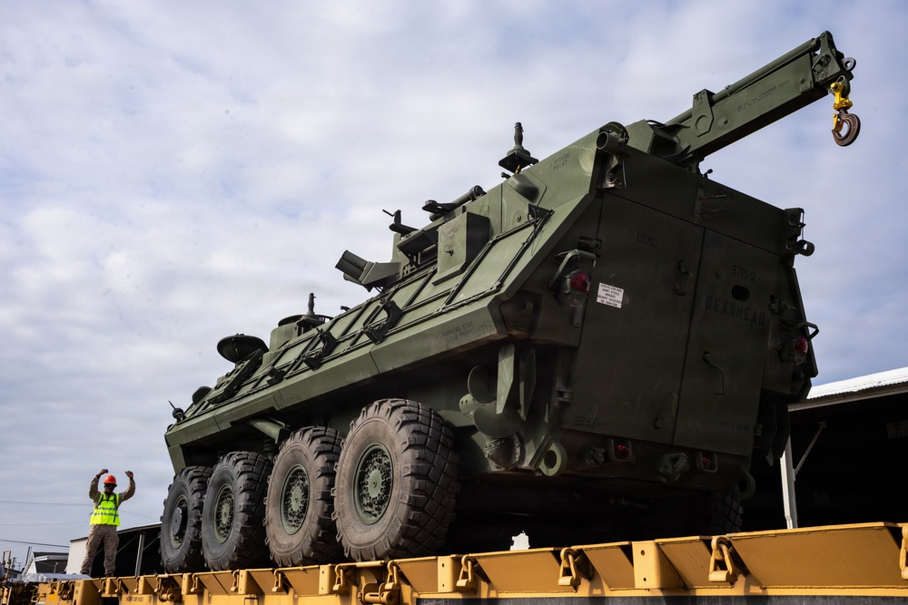 A large olive-drab military armored recovery vehicle, consistent with a Stryker Recovery Vehicle (M1132 or similar variant), is loaded on a yellow flatbed railcar. A worker wearing a high-visibility yellow vest and red hard hat stands beside the vehicle, appearing to signal or guide the loading operation. The scene appears to be at a rail yard or military logistics facility.