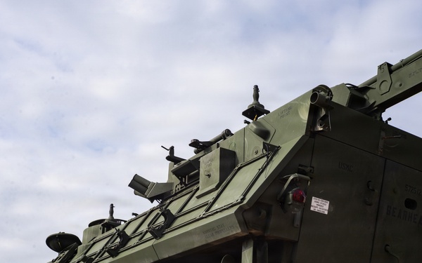 A large olive-drab military armored recovery vehicle, consistent with a Stryker Recovery Vehicle (M1132 or similar variant), is loaded on a yellow flatbed railcar. A worker wearing a high-visibility yellow vest and red hard hat stands beside the vehicle, appearing to signal or guide the loading operation. The scene appears to be at a rail yard or military logistics facility.