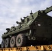A large olive-drab military armored recovery vehicle, consistent with a Stryker Recovery Vehicle (M1132 or similar variant), is loaded on a yellow flatbed railcar. A worker wearing a high-visibility yellow vest and red hard hat stands beside the vehicle, appearing to signal or guide the loading operation. The scene appears to be at a rail yard or military logistics facility.