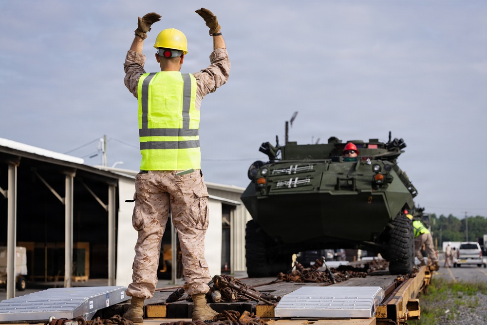 A military personnel wearing a high-visibility safety vest and yellow hard hat is directing a large green armored military vehicle onto a flatbed railcar or transport platform. The scene appears to be a military logistics or equipment loading operation at what looks like a rail yard or depot facility. Additional personnel in safety gear are visible assisting with the operation.