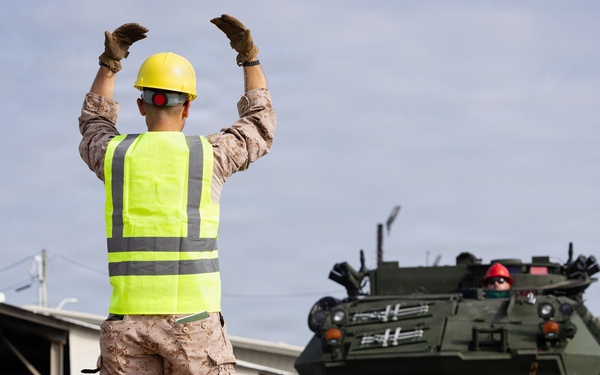 A military personnel wearing a high-visibility safety vest and yellow hard hat is directing a large green armored military vehicle onto a flatbed railcar or transport platform. The scene appears to be a military logistics or equipment loading operation at what looks like a rail yard or depot facility. Additional personnel in safety gear are visible assisting with the operation.