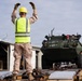 A military personnel wearing a high-visibility safety vest and yellow hard hat is directing a large green armored military vehicle onto a flatbed railcar or transport platform. The scene appears to be a military logistics or equipment loading operation at what looks like a rail yard or depot facility. Additional personnel in safety gear are visible assisting with the operation.