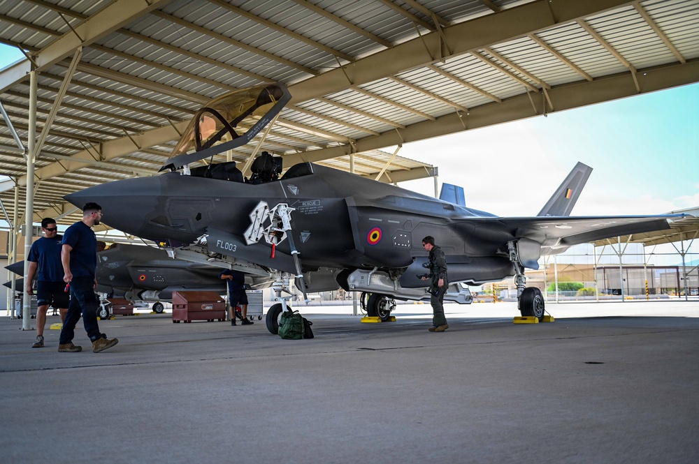 A Belgian Air Component F-35A Lightning II stealth fighter jet (serial number FL003) is parked under a covered hangar, likely at Luke Air Force Base in Arizona. The aircraft displays Belgian roundel markings and what appears to be a special commemorative nose art. Ground crew and a flight-suited pilot are conducting pre-flight or post-flight inspection activities around the aircraft.
