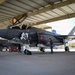 A Belgian Air Component F-35A Lightning II stealth fighter jet (serial number FL003) is parked under a covered hangar, likely at Luke Air Force Base in Arizona. The aircraft displays Belgian roundel markings and what appears to be a special commemorative nose art. Ground crew and a flight-suited pilot are conducting pre-flight or post-flight inspection activities around the aircraft.