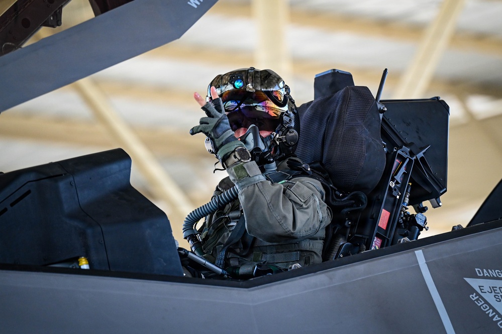 A military pilot is seated in the cockpit of what appears to be an F-35 Lightning II stealth fighter jet, likely inside a hangar. The pilot is wearing a full flight suit, tactical gloves, oxygen mask, and an advanced helmet-mounted display system with a reflective visor. The pilot appears to be giving a thumbs-up gesture toward the camera.