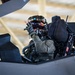 A military pilot is seated in the cockpit of what appears to be an F-35 Lightning II stealth fighter jet, likely inside a hangar. The pilot is wearing a full flight suit, tactical gloves, oxygen mask, and an advanced helmet-mounted display system with a reflective visor. The pilot appears to be giving a thumbs-up gesture toward the camera.