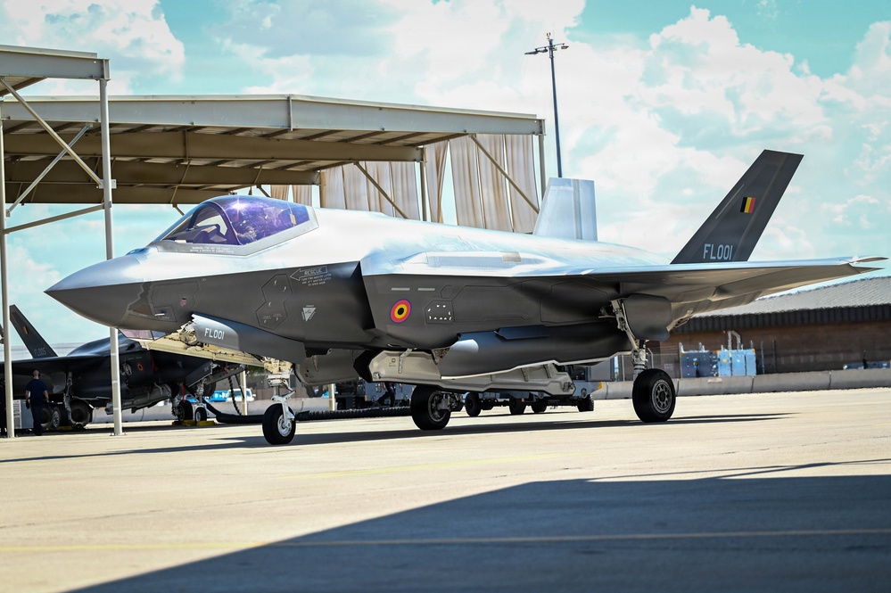 A Belgian Air Component F-35A Lightning II stealth fighter jet, serial number FL001, is parked on an airfield apron. The aircraft displays the Belgian national roundel (black, yellow, red) on its fuselage and tail fin. It appears to be in a static ground display or pre-flight configuration near a hangar structure.