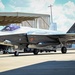 A Belgian Air Component F-35A Lightning II stealth fighter jet, serial number FL001, is parked on an airfield apron. The aircraft displays the Belgian national roundel (black, yellow, red) on its fuselage and tail fin. It appears to be in a static ground display or pre-flight configuration near a hangar structure.