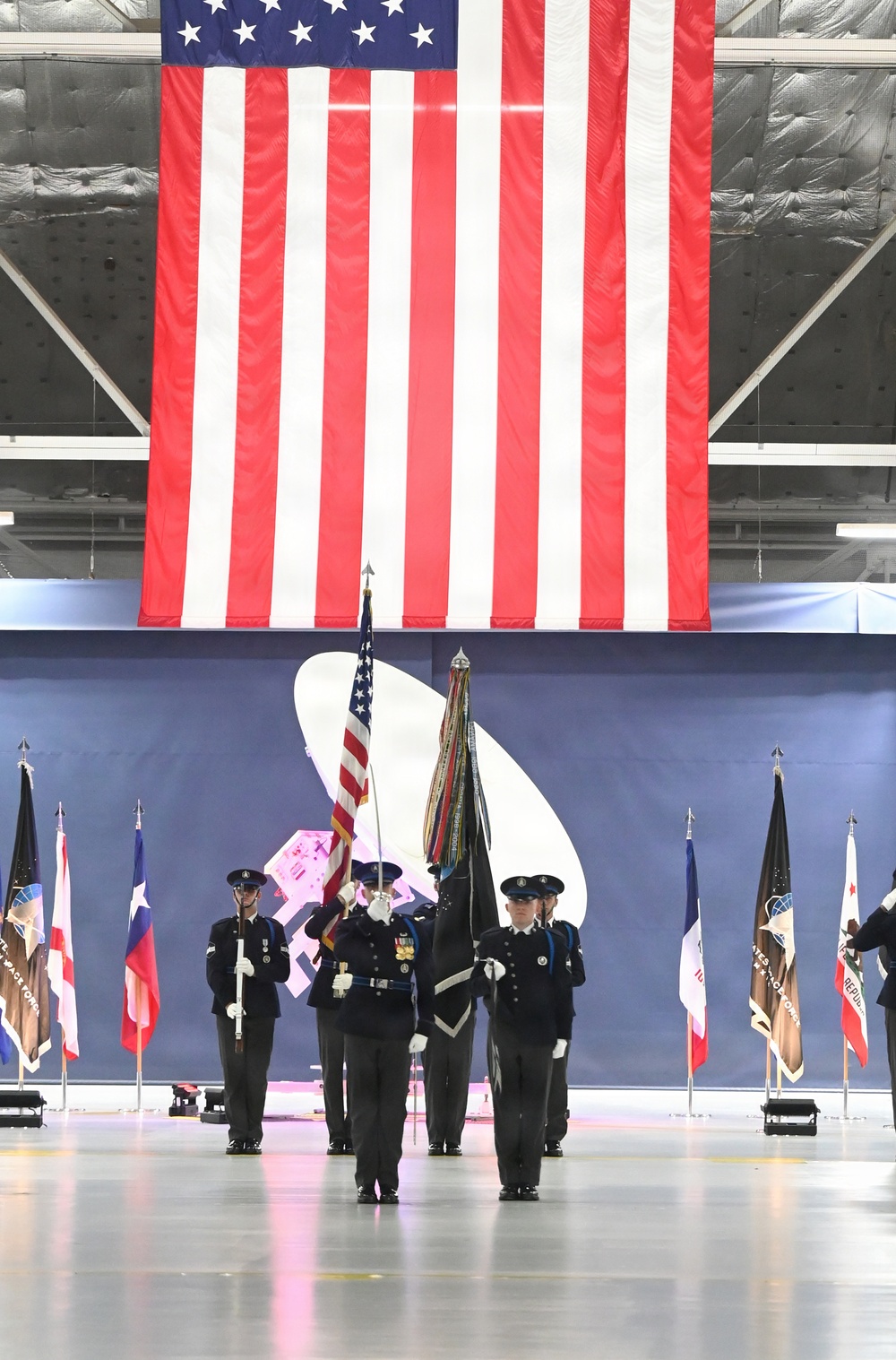 A military color guard ceremony is taking place inside a large hangar or indoor venue. Personnel in dark dress uniforms are presenting colors beneath an oversized American flag hanging from the ceiling. State and organizational flags are displayed on stands flanking the color guard formation.