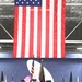A military color guard ceremony is taking place inside a large hangar or indoor venue. Personnel in dark dress uniforms are presenting colors beneath an oversized American flag hanging from the ceiling. State and organizational flags are displayed on stands flanking the color guard formation.