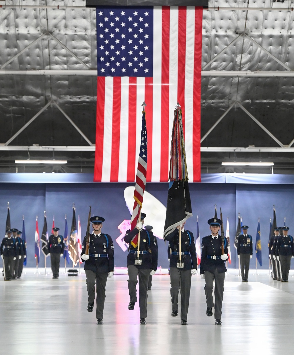 A formal military color guard ceremony is taking place inside a large aircraft hangar. Four uniformed service members march in formation carrying the U.S. national flag and a unit guidon with streamers, while additional personnel line the sides holding various organizational flags. A large American flag is prominently displayed hanging from the hangar's rear wall as a backdrop.