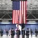 A formal military color guard ceremony is taking place inside a large aircraft hangar. Four uniformed service members march in formation carrying the U.S. national flag and a unit guidon with streamers, while additional personnel line the sides holding various organizational flags. A large American flag is prominently displayed hanging from the hangar's rear wall as a backdrop.