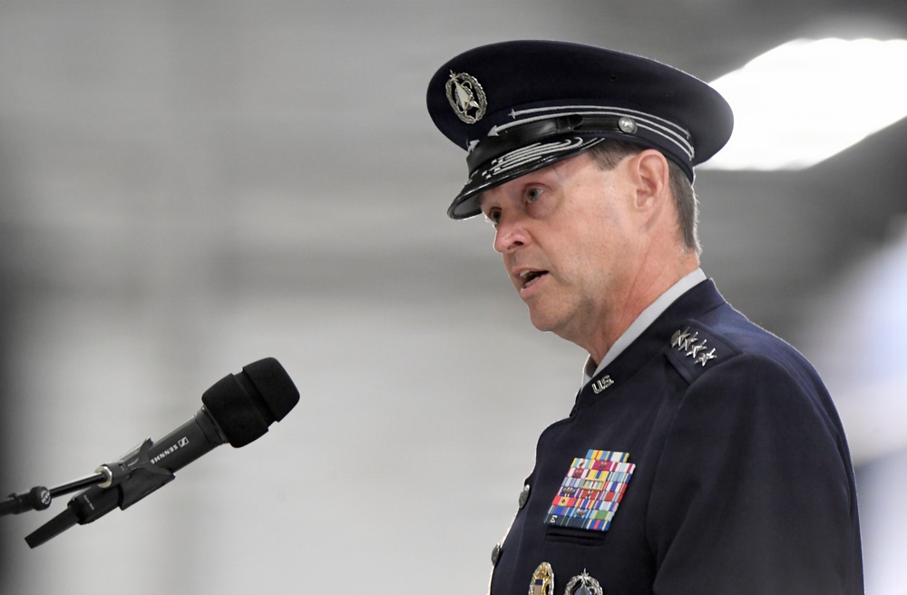 A senior U.S. Air Force officer in dress uniform is speaking at a podium with a Sennheiser microphone. The officer displays multiple rows of service ribbons, medals, and rank insignia consistent with a high-ranking general officer. The setting appears to be an indoor military ceremony or official event.