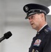 A senior U.S. Air Force officer in dress uniform is speaking at a podium with a Sennheiser microphone. The officer displays multiple rows of service ribbons, medals, and rank insignia consistent with a high-ranking general officer. The setting appears to be an indoor military ceremony or official event.