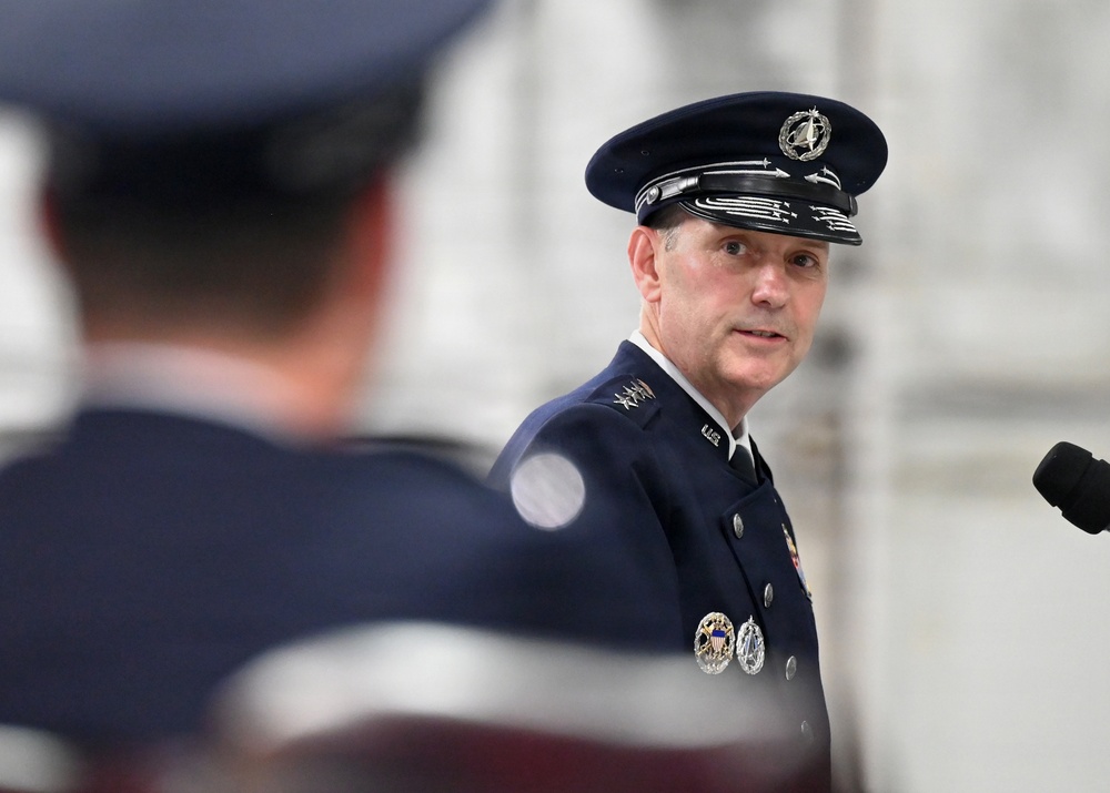 A senior military officer in dress uniform is speaking at what appears to be a formal military ceremony. The officer wears a dark blue service dress uniform with multiple decorations and a peaked cap featuring a distinctive emblem. A second uniformed individual is visible in the blurred foreground.