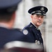A senior military officer in dress uniform is speaking at what appears to be a formal military ceremony. The officer wears a dark blue service dress uniform with multiple decorations and a peaked cap featuring a distinctive emblem. A second uniformed individual is visible in the blurred foreground.