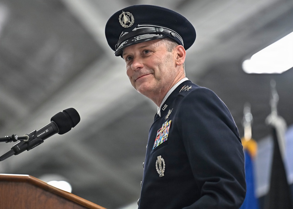 A senior U.S. Space Force or Air Force officer in dress uniform stands at a podium with a Sennheiser microphone, appearing to deliver a speech or remarks at an official military ceremony. The officer wears a dark blue service dress uniform adorned with multiple ribbons, badges, and rank insignia consistent with a high-ranking general officer. The setting appears to be an indoor military facility or hangar with flags visible in the background.
