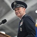 A senior U.S. Space Force or Air Force officer in dress uniform stands at a podium with a Sennheiser microphone, appearing to deliver a speech or remarks at an official military ceremony. The officer wears a dark blue service dress uniform adorned with multiple ribbons, badges, and rank insignia consistent with a high-ranking general officer. The setting appears to be an indoor military facility or hangar with flags visible in the background.