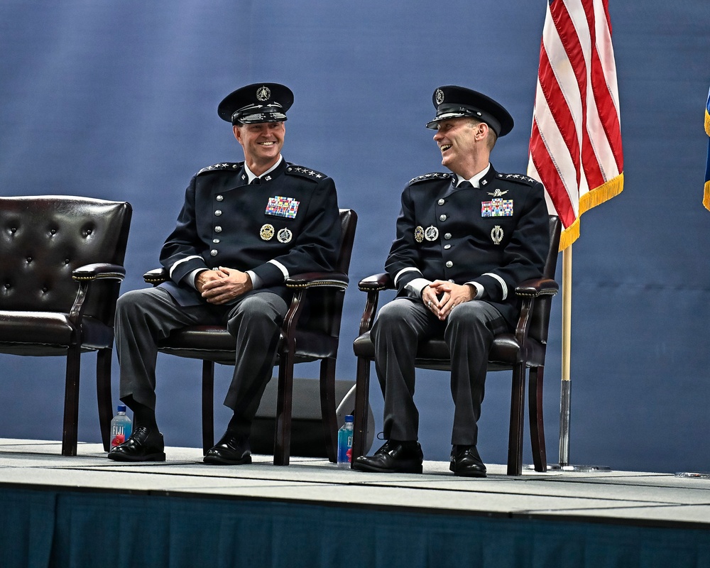 Two senior military officers in dark dress uniforms are seated on a stage, sharing a laugh during what appears to be a formal military ceremony. Both wear high-ranking insignia consistent with general officer grades, multiple service ribbons, and distinctive unit badges. An American flag is visible to their right, and the setting suggests an official change of command or awards ceremony.