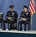 Two senior military officers in dark dress uniforms are seated on a stage, sharing a laugh during what appears to be a formal military ceremony. Both wear high-ranking insignia consistent with general officer grades, multiple service ribbons, and distinctive unit badges. An American flag is visible to their right, and the setting suggests an official change of command or awards ceremony.