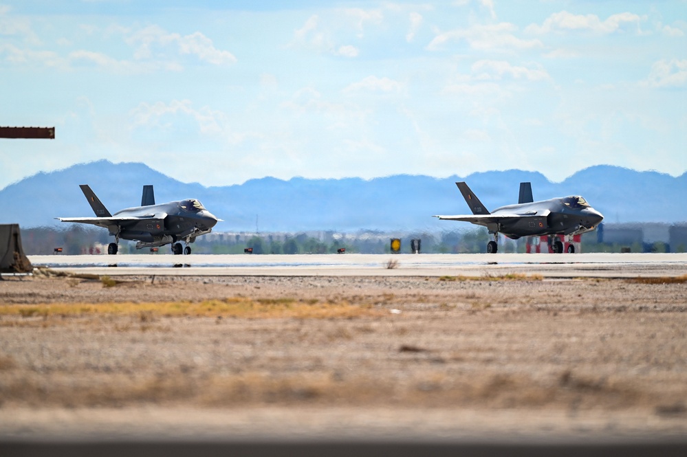Two F-35 Lightning II stealth fighter jets are taxiing on a runway at what appears to be a desert military airbase. The aircraft are positioned side by side on the tarmac with arid terrain and mountain ranges visible in the background. The setting suggests a southwestern United States location based on the desert landscape and terrain features.