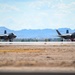 Two F-35 Lightning II stealth fighter jets are taxiing on a runway at what appears to be a desert military airbase. The aircraft are positioned side by side on the tarmac with arid terrain and mountain ranges visible in the background. The setting suggests a southwestern United States location based on the desert landscape and terrain features.