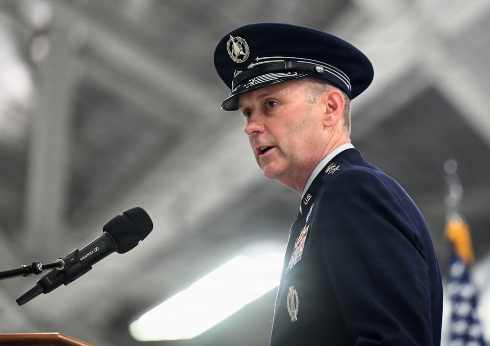 A senior United States Air Force officer in dress uniform is speaking at a podium with a Sennheiser microphone. The officer is wearing a service dress cap with scrambled eggs insignia and a heavily decorated uniform with multiple ribbons and badges. The setting appears to be an official military ceremony held inside a large hangar or similar facility.