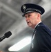 A senior United States Air Force officer in dress uniform is speaking at a podium with a Sennheiser microphone. The officer is wearing a service dress cap with scrambled eggs insignia and a heavily decorated uniform with multiple ribbons and badges. The setting appears to be an official military ceremony held inside a large hangar or similar facility.