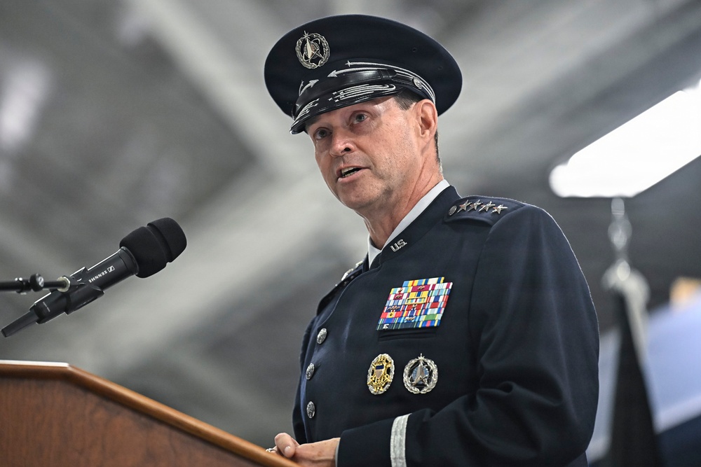 A senior military officer in dress uniform is speaking at a podium with a Sennheiser microphone. The uniform features four stars on the shoulder, indicating the rank of General, along with numerous service ribbons, badges, and a cap bearing what appears to be a Space Force or Air Force emblem. The setting appears to be an indoor military facility such as a hangar.