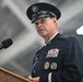 A senior military officer in dress uniform is speaking at a podium with a Sennheiser microphone. The uniform features four stars on the shoulder, indicating the rank of General, along with numerous service ribbons, badges, and a cap bearing what appears to be a Space Force or Air Force emblem. The setting appears to be an indoor military facility such as a hangar.