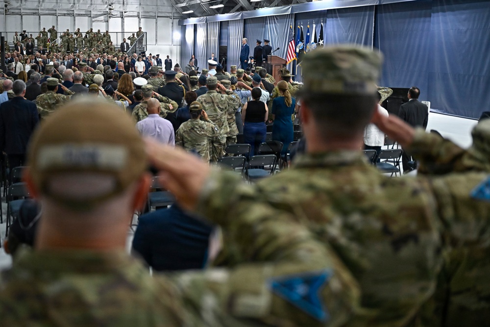 A large military ceremony is taking place inside what appears to be a hangar or large indoor facility. Numerous military personnel in camouflage uniforms are rendering salutes toward a stage or podium area at the front, where officials and flags are visible. The event appears to be a formal military ceremony such as a change of command, promotion, or official address.