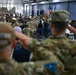 A large military ceremony is taking place inside what appears to be a hangar or large indoor facility. Numerous military personnel in camouflage uniforms are rendering salutes toward a stage or podium area at the front, where officials and flags are visible. The event appears to be a formal military ceremony such as a change of command, promotion, or official address.