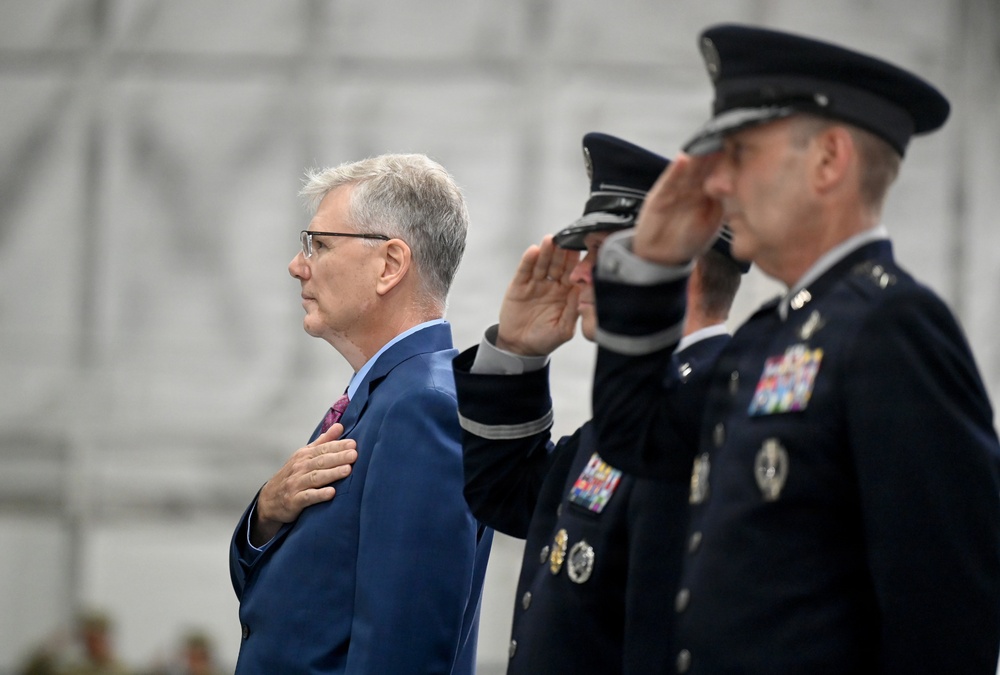 The image shows a formal military or official ceremony with individuals rendering honors, likely during a national anthem or flag presentation. A civilian in a blue suit places his hand over his heart while military officers in dark blue uniforms salute. The setting appears to be inside a large hangar or tent structure.
