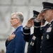 The image shows a formal military or official ceremony with individuals rendering honors, likely during a national anthem or flag presentation. A civilian in a blue suit places his hand over his heart while military officers in dark blue uniforms salute. The setting appears to be inside a large hangar or tent structure.