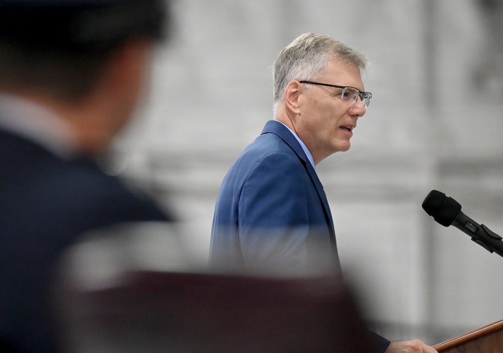 A middle-aged man with gray hair and glasses is speaking at a podium with a microphone. He is wearing a blue blazer and appears to be delivering a formal address or speech. In the foreground, a blurred figure wearing a dark uniform cap is partially visible.