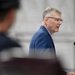 A middle-aged man with gray hair and glasses is speaking at a podium with a microphone. He is wearing a blue blazer and appears to be delivering a formal address or speech. In the foreground, a blurred figure wearing a dark uniform cap is partially visible.