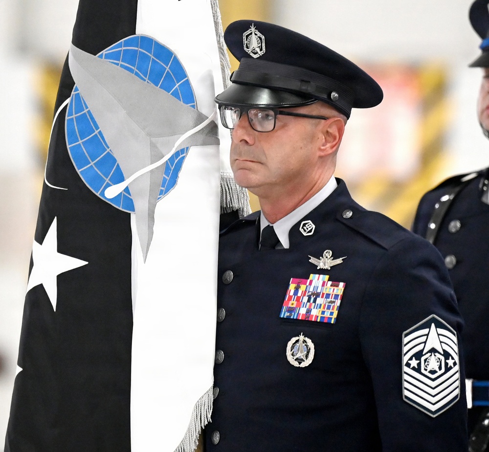 A senior non-commissioned officer in dress uniform stands at attention during a formal military ceremony, holding a unit guidon or organizational flag. The individual displays the rank insignia of a Chief Master Sergeant of the Space Force or Air Force, along with numerous service ribbons and badges. The flag features a globe, star, and spacecraft imagery consistent with U.S. Space Force or Air Mobility Command heraldry.