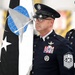 A senior non-commissioned officer in dress uniform stands at attention during a formal military ceremony, holding a unit guidon or organizational flag. The individual displays the rank insignia of a Chief Master Sergeant of the Space Force or Air Force, along with numerous service ribbons and badges. The flag features a globe, star, and spacecraft imagery consistent with U.S. Space Force or Air Mobility Command heraldry.