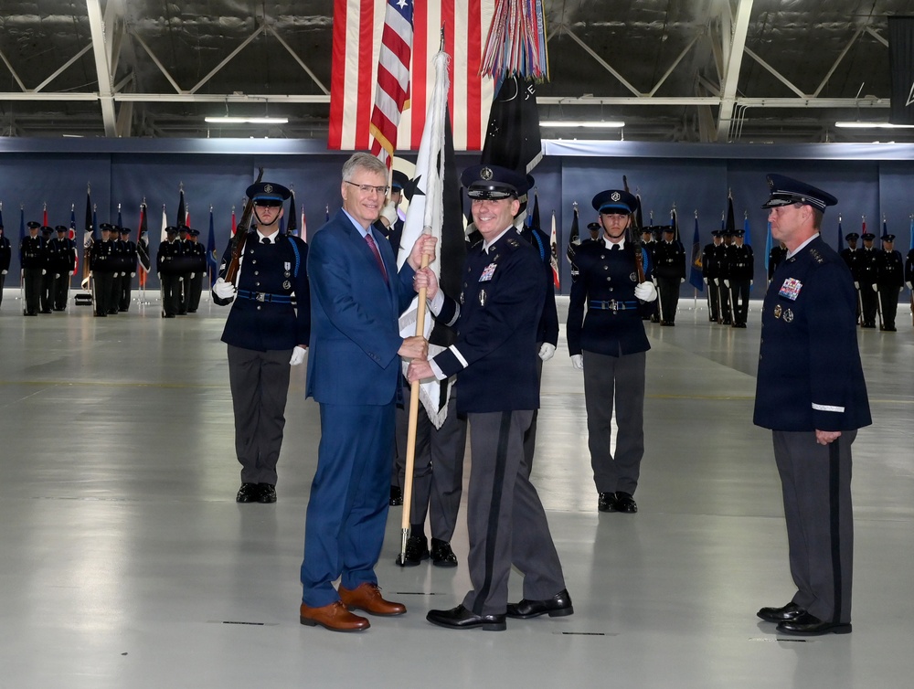 A formal military change of command or activation ceremony taking place inside a large hangar or indoor venue. A civilian in a blue suit and a uniformed military officer are jointly holding a guidon/unit flag, which is the central symbolic act of such ceremonies. Honor guard members stand at attention in the background alongside multiple national and unit flags.