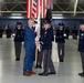 A formal military change of command or activation ceremony taking place inside a large hangar or indoor venue. A civilian in a blue suit and a uniformed military officer are jointly holding a guidon/unit flag, which is the central symbolic act of such ceremonies. Honor guard members stand at attention in the background alongside multiple national and unit flags.