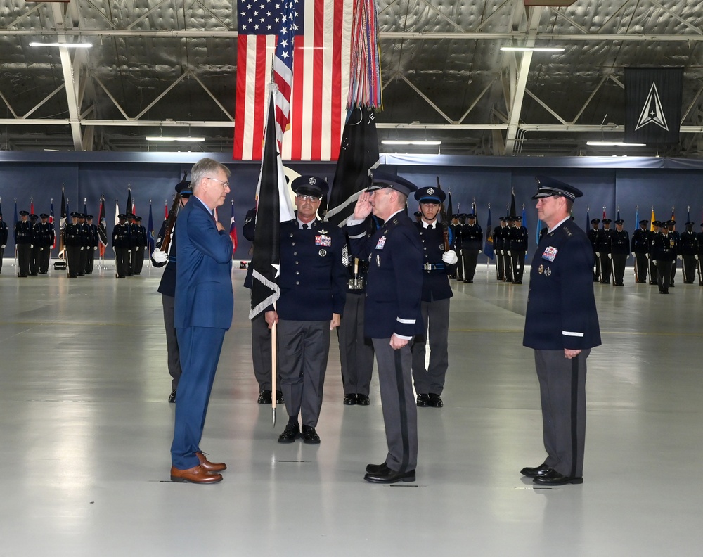 A formal military ceremony is taking place inside a large hangar facility, likely a change of command or activation ceremony. A civilian in a blue suit faces uniformed military officers, one of whom is rendering a salute, while a guidon bearer holds a dark ceremonial flag. Rows of uniformed personnel stand in formation in the background beneath a large American flag and what appears to be a U.S. Space Force delta emblem.