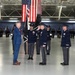A formal military ceremony is taking place inside a large hangar facility, likely a change of command or activation ceremony. A civilian in a blue suit faces uniformed military officers, one of whom is rendering a salute, while a guidon bearer holds a dark ceremonial flag. Rows of uniformed personnel stand in formation in the background beneath a large American flag and what appears to be a U.S. Space Force delta emblem.