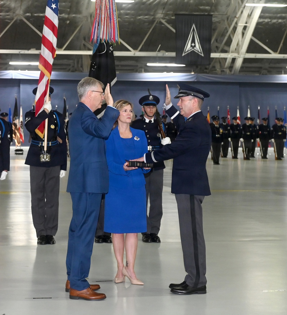 A formal military swearing-in ceremony taking place inside a large hangar or ceremonial hall. A civilian in a blue suit is raising his right hand while a uniformed military officer administers the oath, with a woman in a blue dress holding a Bible between them. The background features honor guard personnel, multiple flags including the U.S. flag, and a U.S. Space Force emblem visible on the wall.