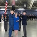A formal military swearing-in ceremony taking place inside a large hangar or ceremonial hall. A civilian in a blue suit is raising his right hand while a uniformed military officer administers the oath, with a woman in a blue dress holding a Bible between them. The background features honor guard personnel, multiple flags including the U.S. flag, and a U.S. Space Force emblem visible on the wall.