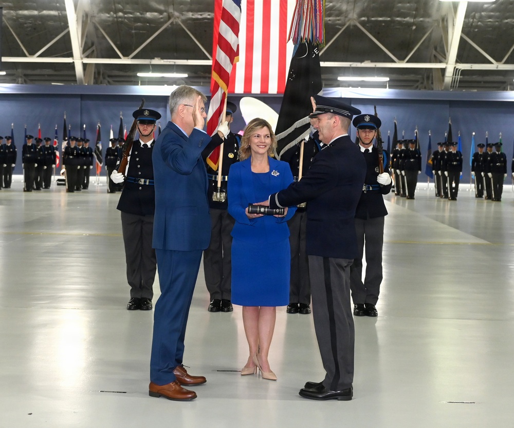 A formal military swearing-in or commissioning ceremony taking place inside what appears to be a large aircraft hangar. A civilian in a blue suit is rendering a salute while a uniformed military officer administers an oath, with a woman in a blue dress holding what appears to be a Bible or book. Honor guard members in dark dress uniforms with rifles are lined up in formation in the background alongside multiple flags.